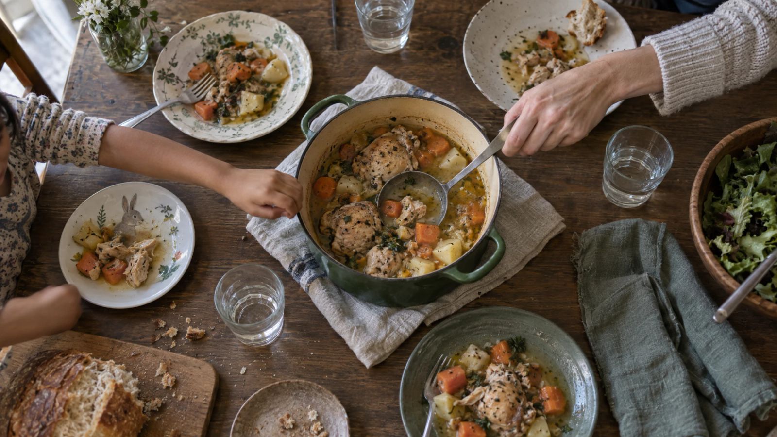 A family dinner table from above with four plates and two hands, a child's and an adult's, reaching for the same pot.